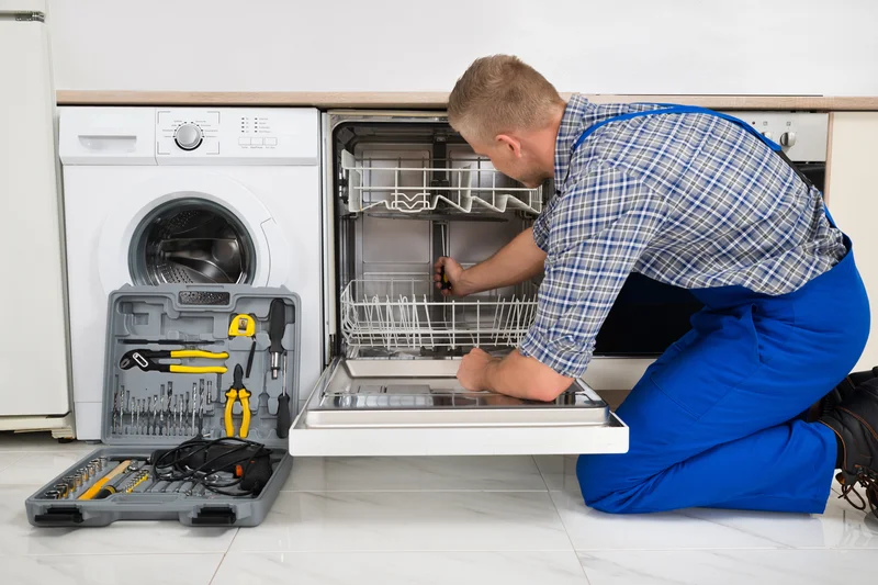 Technician in kitchen repairing open dishwasher near washing machine.